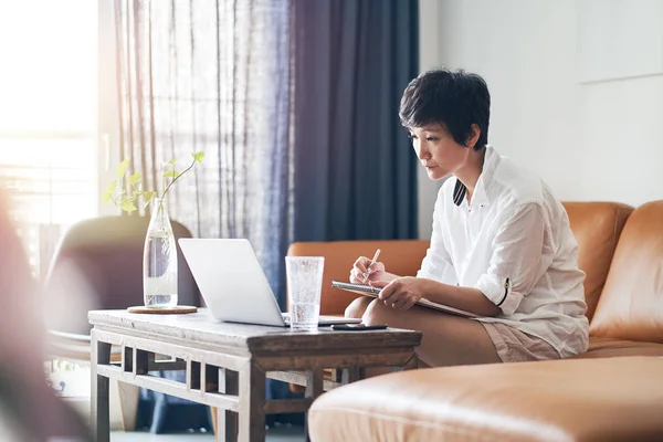 Asyalı bir kadın evdeki kanepede oturup dizüstü bilgisayarla çalışıyor.