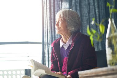 Yaşlı Asyalı kadın evde kitap okuyor emekliliğin tadını çıkarıyor.