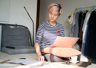 Güzel Asyalı kadın yatakta oturuyor ve dizüstü bilgisayarla evden çalışıyor