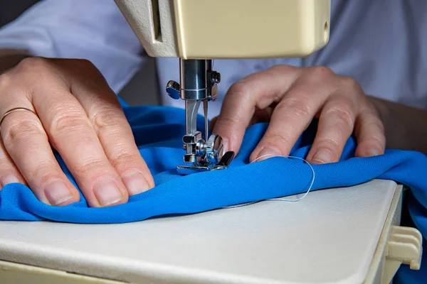 Female hands sew a blue dress on a professional overlock machine in a workshop. Seamstress hands holding textile for sewing clothes. Blurred background Close-up view of the sewing process. — Stock Photo, Image