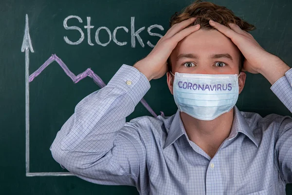 Young businessman in a medical mask with the inscription coronavirus, clutching his head in horror, in the background is a stock chart with a negative trend. Coronavirus and prevention of financial