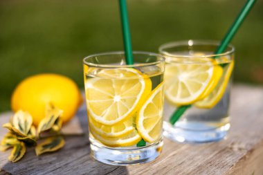 Yaz içeceği, limon dilimleriyle dolu bir bardak su, güneşli sıcak bir günde çimenlikte tahta bir tahta üzerinde durmak, arka plan bulanık, sığ bir alan derinliği, seçici bir odak noktası..