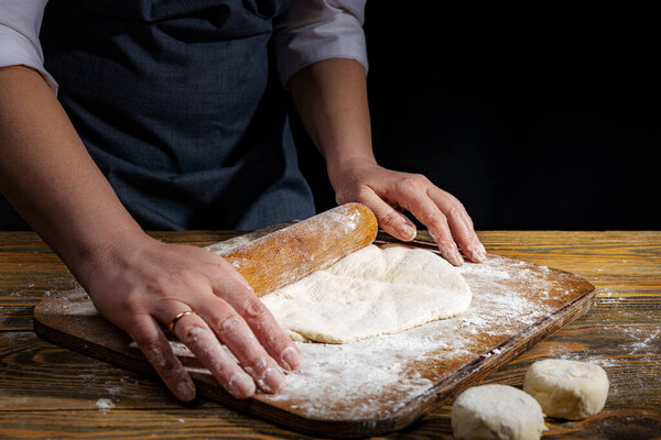 Female hands knead the dough on a wooden antique table on a dark background, close-up, shallow depth of field, beautiful directional lighting. Concept of home baking and comfort.
