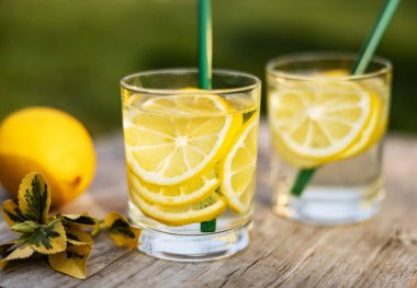 Yaz içeceği, limon dilimleriyle dolu bir bardak su, güneşli sıcak bir günde çimenlikte tahta bir tahta üzerinde durmak, arka plan bulanık, sığ bir alan derinliği, seçici bir odak noktası..