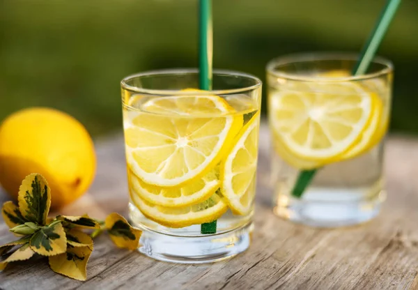 Yaz içeceği, limon dilimleriyle dolu bir bardak su, güneşli sıcak bir günde çimenlikte tahta bir tahta üzerinde durmak, arka plan bulanık, sığ bir alan derinliği, seçici bir odak noktası..