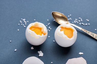 Mavi pastel arka planda kaynamış yumurta, yarısı soyulmuş, yanında kabuk, tuz ve bir kaşık var. Hızlı ve kolay kahvaltı konsepti.