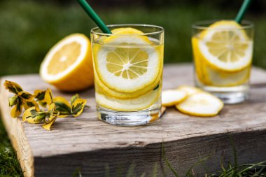 Yaz içeceği, limon dilimleriyle dolu bir bardak su, güneşli sıcak bir günde çimenlikte tahta bir tahta üzerinde durmak, arka plan bulanık, sığ bir alan derinliği, seçici bir odak noktası..