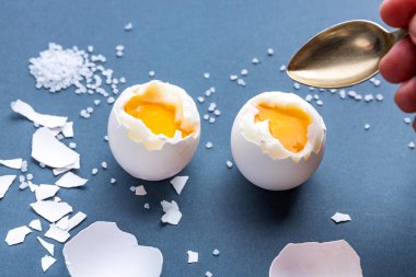 Mavi pastel arka planda kaynamış yumurta, yarısı soyulmuş, yanında kabuk, ekmek, tuz ve mayonez, bir el kaşık tutuyor. Hızlı ve kolay kahvaltı konsepti.