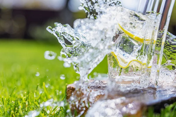 Une boisson d'été, un verre d'eau avec des éclaboussures remplies de tranches de citrons et de glace, debout sur une planche de bois sur la pelouse par une journée chaude ensoleillée, le fond est flou, peu profonde profondeur de champ — Photo