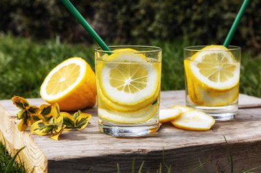 Yaz içeceği, limon dilimleriyle dolu bir bardak su, güneşli sıcak bir günde çimenlikte tahta bir tahta üzerinde durmak, arka plan bulanık, sığ bir alan derinliği, seçici bir odak noktası..
