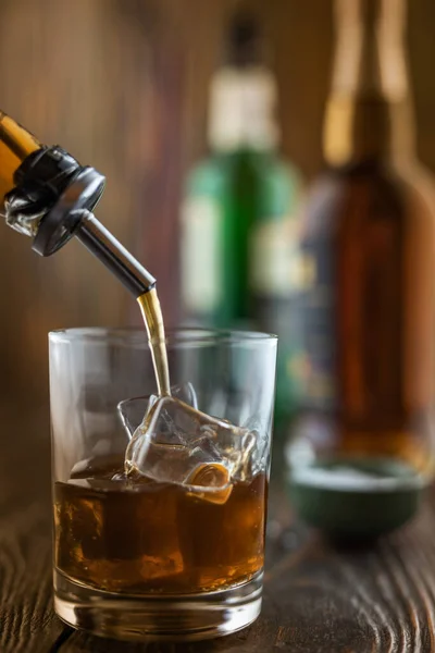 In the bar, whiskey is poured into a glass with ice, in the background there are bottles on a wooden table of the bar counter, shallow depth of field, selective focus. The concept of alcoholic drinks