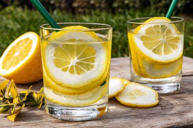 Yaz içeceği, limon dilimleriyle dolu bir bardak su, güneşli sıcak bir günde çimenlikte tahta bir tahta üzerinde durmak, arka plan bulanık, sığ bir alan derinliği, seçici bir odak noktası..