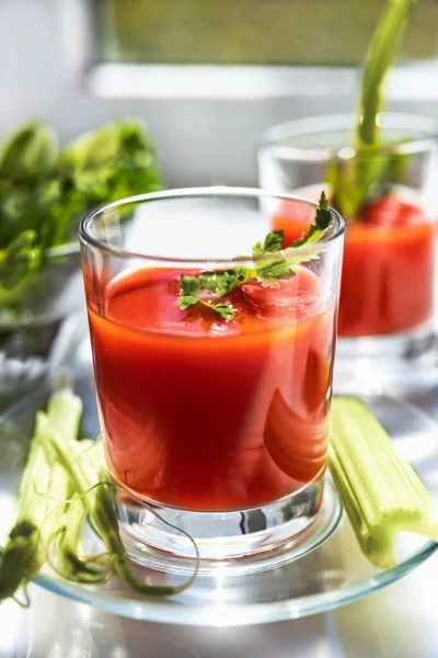 Dos vasos de jugo de tomate con perejil y decoraciones de apio, de pie cerca de la ventana, la luz del sol de la mañana brilla, poca profundidad de campo, enfoque selectivo. Concepto de bebidas naturales . — Foto de Stock