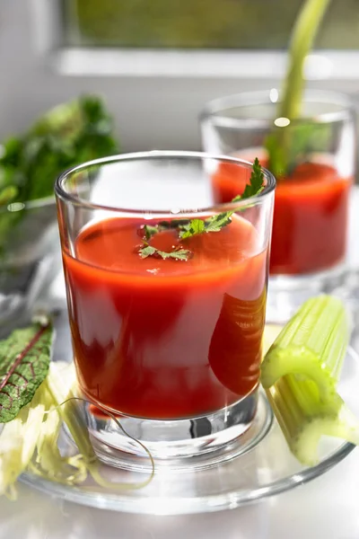 Dos vasos de jugo de tomate con perejil y decoraciones de apio, de pie cerca de la ventana, la luz del sol de la mañana brilla, poca profundidad de campo, enfoque selectivo. Concepto de bebidas naturales . — Foto de Stock