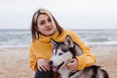Sarı kazaklı genç bir kadın kumsalda bir köpekle oynar (gri ve beyaz husky)