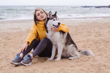 Sarı kazaklı genç bir kadın kumsalda bir köpekle oynar (gri ve beyaz husky)