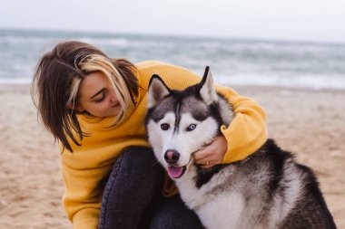 Sarı kazaklı genç bir kadın kumsalda bir köpekle oynar (gri ve beyaz husky)