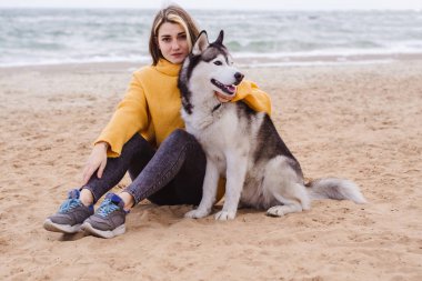 Sarı kazaklı genç bir kadın kumsalda bir köpekle oynar (gri ve beyaz husky)