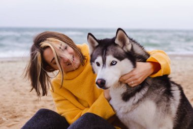 Sarı kazaklı genç bir kadın kumsalda bir köpekle oynar (gri ve beyaz husky)