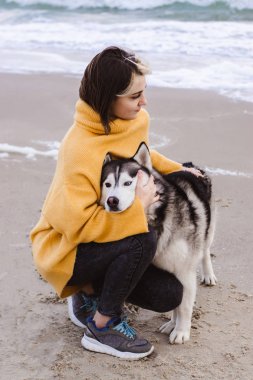 Sarı kazaklı genç bir kadın kumsalda bir köpekle oynar (gri ve beyaz husky)