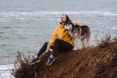 Sarı kazaklı genç bir kadın kumsalda bir köpekle oynar (gri ve beyaz husky)
