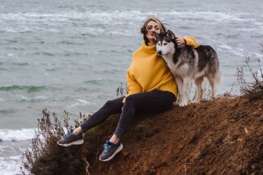 Sarı kazaklı genç bir kadın kumsalda bir köpekle oynar (gri ve beyaz husky)