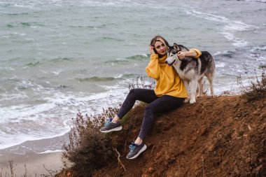 Sarı kazaklı genç bir kadın kumsalda bir köpekle oynar (gri ve beyaz husky)