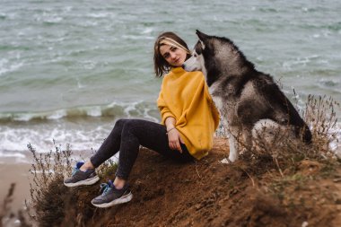 Sarı kazaklı genç bir kadın kumsalda bir köpekle oynar (gri ve beyaz husky)