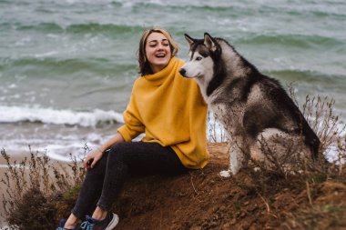 Sarı kazaklı genç bir kadın kumsalda bir köpekle oynar (gri ve beyaz husky)
