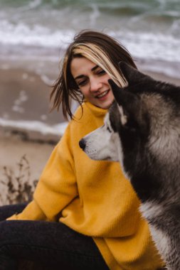Sarı kazaklı genç bir kadın kumsalda bir köpekle oynar (gri ve beyaz husky)