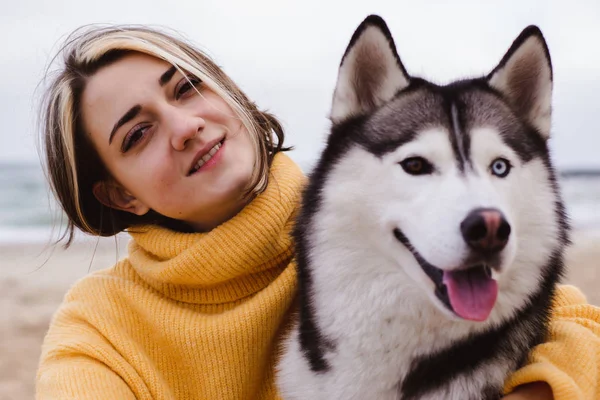 Sarı kazaklı genç bir kadın kumsalda bir köpekle oynar (gri ve beyaz husky)