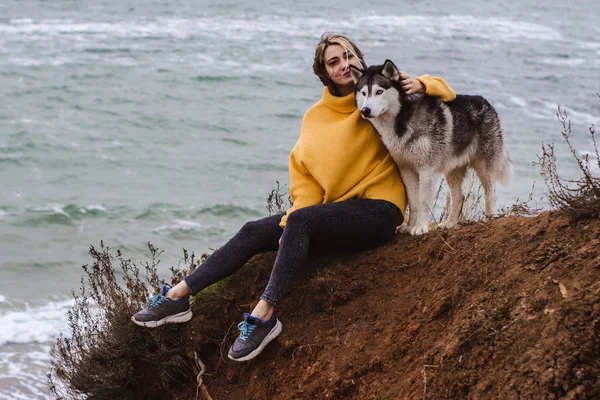 Sarı kazaklı genç bir kadın kumsalda bir köpekle oynar (gri ve beyaz husky)