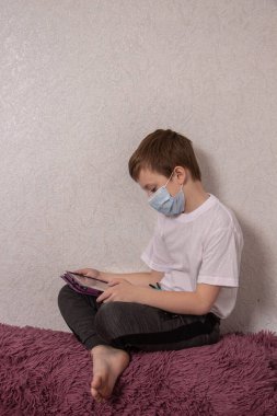 Tıbbi maske takan zeki bir çocuk kendini koronavirüsten korur ve tablete bakar.
