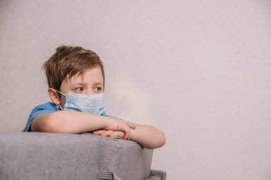 Mavi tişörtlü, maske takmış üzgün bir bebek kanepede tek başına oturuyor.