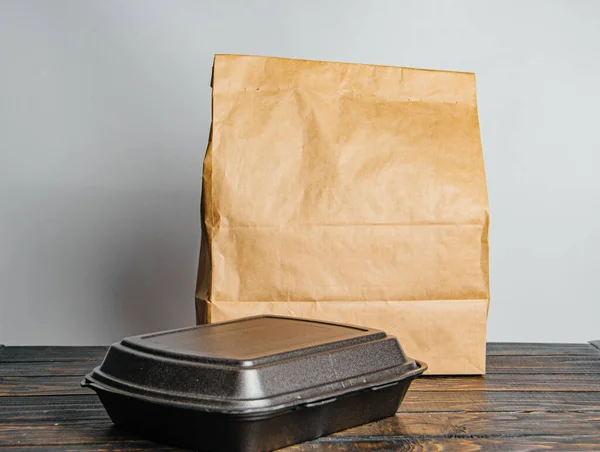 Um saco de papel marrom artesanal e um recipiente preto para tirar ou entregar mercadorias e alimentos em uma mesa de madeira. Lugar para publicidade. conceito de serviço de entrega — Fotografia de Stock