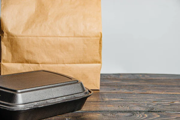 A brown craft paper bag and a black container for taking out or delivering goods and food on a wooden table. Place for advertising. delivery service concept