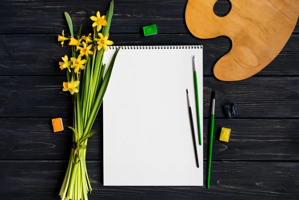 Livro de esboços, aquarela e pincéis em mesa de madeira preta com buquê de narcisos. Deitado plano, vista superior — Fotografia de Stock