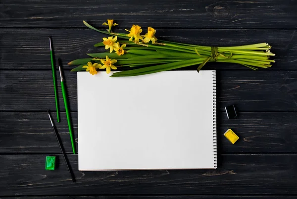 Skicář, akvarel a štětce na černý dřevěný stůl s kytice narcisů. Plochá ležel, horní pohled — Stock fotografie