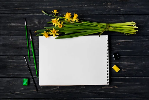 Livro de esboços, aquarela e pincéis em mesa de madeira preta com buquê de narcisos. Deitado plano, vista superior — Fotografia de Stock