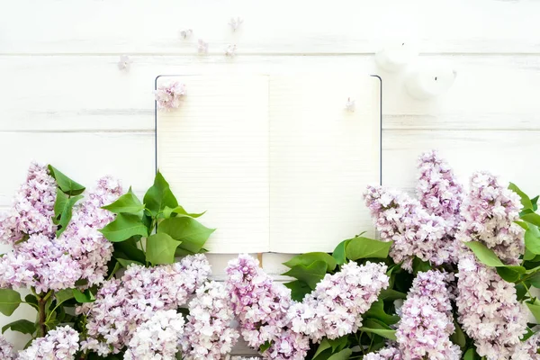 Abra o caderno vazio rodeado de flores lilás sobre fundo branco de madeira. Flat lay, vista superior, espaço de cópia — Fotografia de Stock