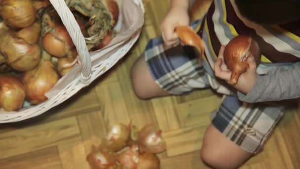 Niño pequeño limpia cebollas sentado en el suelo en la cocina — Vídeo de stock
