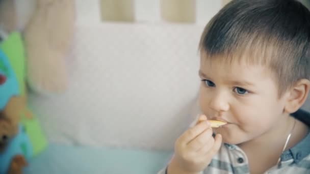 Mały chłopiec siedzi na łóżku i zjada pyszne paluszki słodkie — Wideo stockowe
