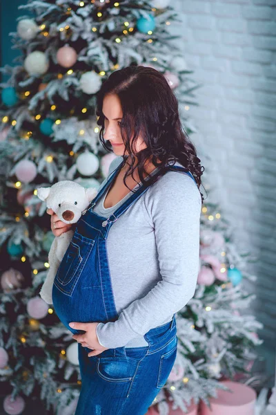 Een mooie zwangere meisje in een overall blauw denim houdt een beer in haar handen en kijkt hem — Stockfoto