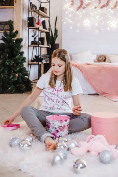 Yeni yıl süslemeleriyle dolu bir odada yerde oturan ve pembe silindirik kutularla, Noel toplarıyla, pembe fareyle ve bir tavşanla oynayan bir kız.