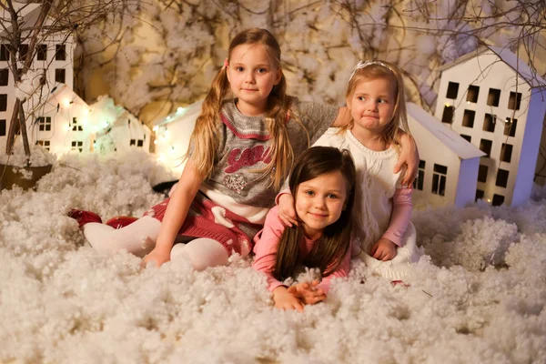 Üç kız, yeni yıl süslemelerinde yapay kar ve küçük karton evlerle sarılıp gülümsüyor.