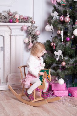 Küçük kız şöminenin yanında tahta ata biniyor ve yılbaşı ağacı