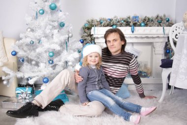 Baba ve kızı süslü şöminenin ve beyaz yapay Noel ağacının yanında oturuyor.