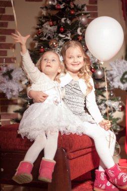 Beyaz ve mor balonlu iki kız Noel süsleri içinde kırmızı bir koltukta oturuyorlar.