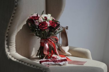 wedding accessories on a beige armchair: a bride's bouquet of red and pink roses, women's shoes, a garter of the bride