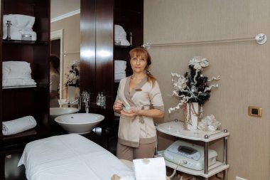 Kadın masaj terapisti masaj masasının yanında duruyor ve prosedürleri uygulamaya hazırlanıyor.